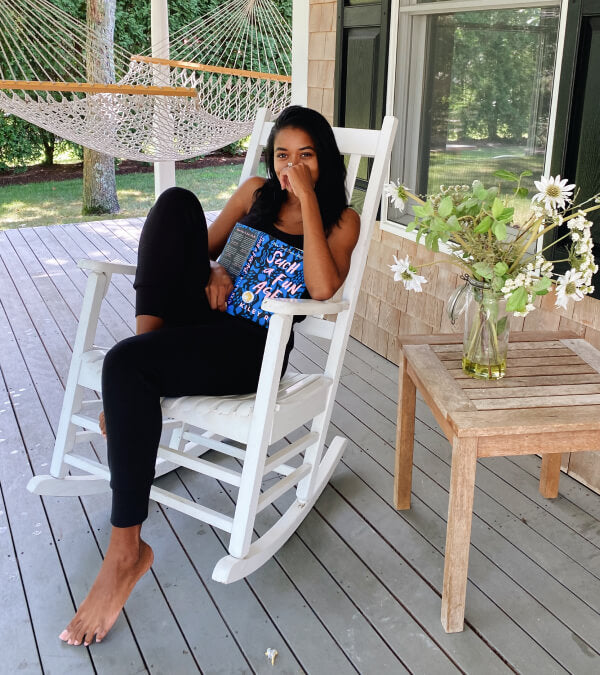 Woman on a porch laughing and holding a book.  Flowers on a table next to her and a hammock in the background