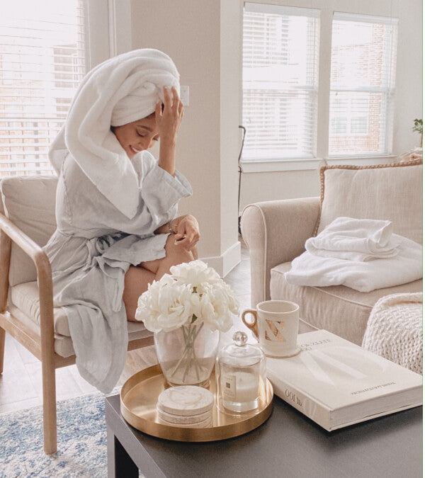 Woman in an all white room in a white linen robe, with her hair twisted up in a white towel, drying.