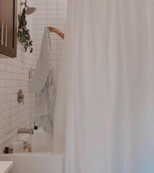 White bathroom with a white shower curtain.  An arm reaches out from the shower holding a white linen robe.