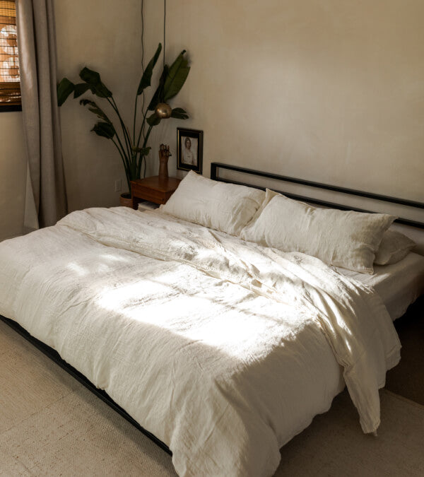 Bed made up in rumpled cream linen sheets.  Everything in the room is beige. Big palm plant in the background