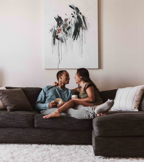 Laught couple in loungewear sitting on a couch in front of a large piece of artwork